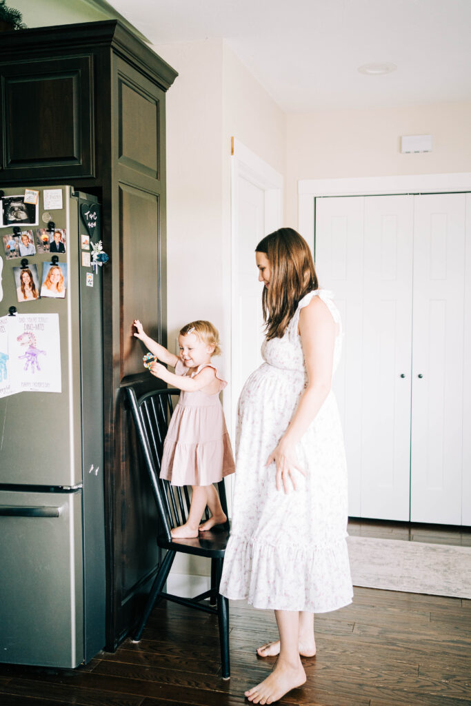 woman pregnant wearing a white floral dress while standing in a kitchen with toddler daughter in a pink dress.