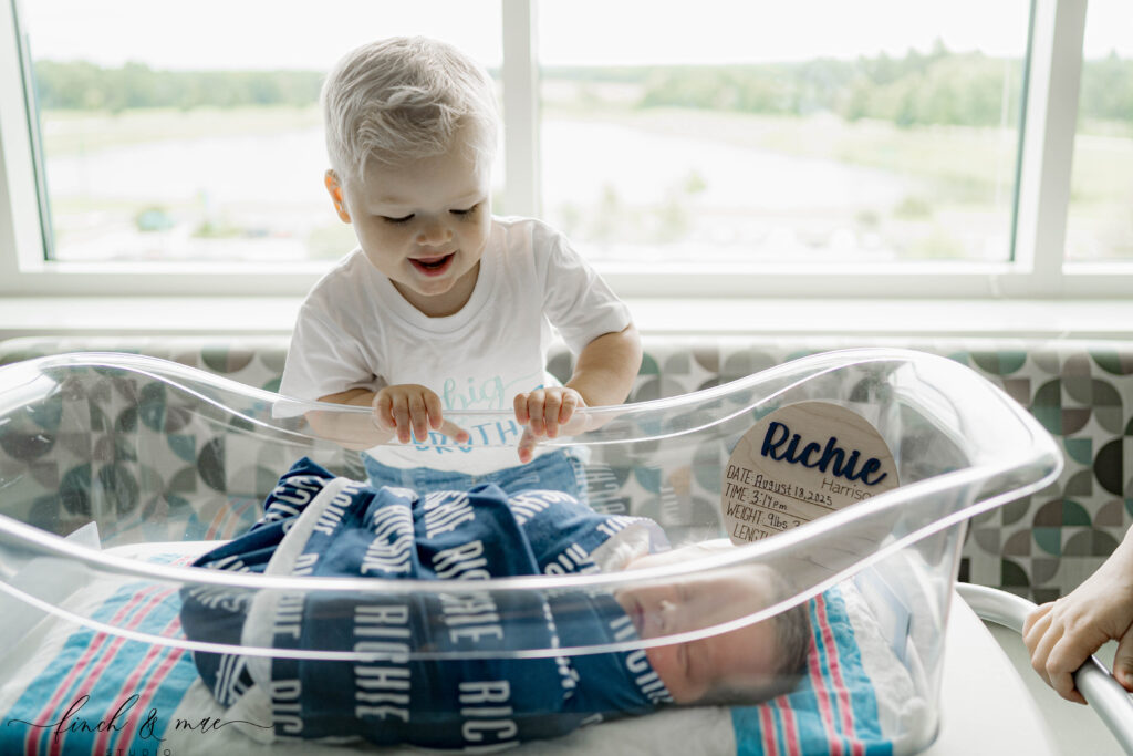 Big brother smiling while meeting his newborn sibling in a hospital bassinet during Fresh 48 photography in Madison, Wisconsin.