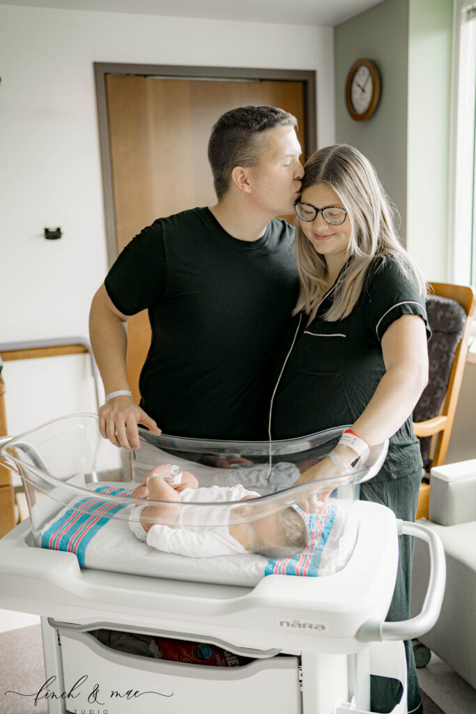 Couple admiring newborn in hospital bassinet – Mom and dad standing together by the bassinet, with dad kissing mom on the forehead while she rests her hand on the baby.