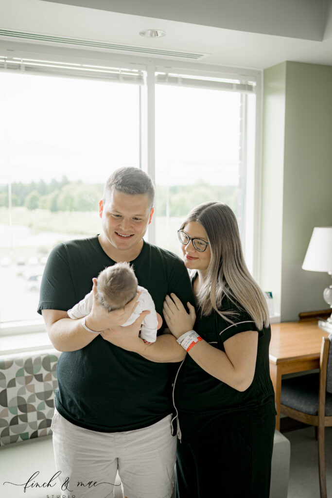 Parents smiling at newborn in hospital room – A father holding the baby while the mother rests her hand on his arm, both smiling down at their newborn.