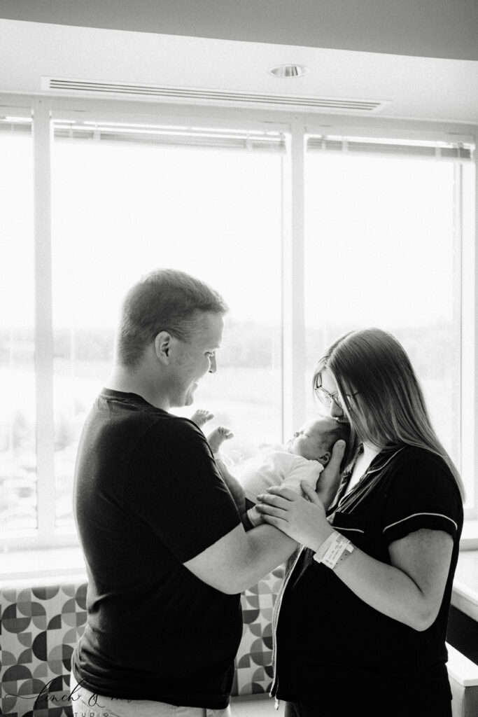 Parents standing together holding newborn baby – Black and white photo of new mom and dad by the hospital window, looking lovingly at their newborn son.