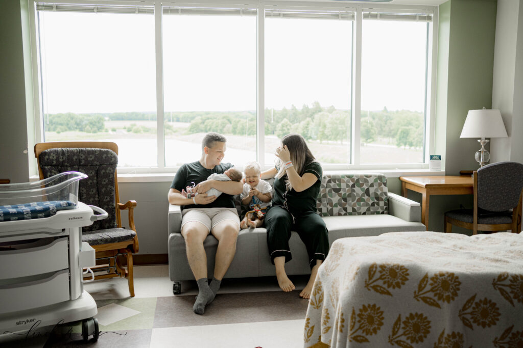 Family sitting together on hospital couch during Fresh 48 session – dad holding newborn baby while mom sits beside him with their toddler, sharing a sweet first sibling moment