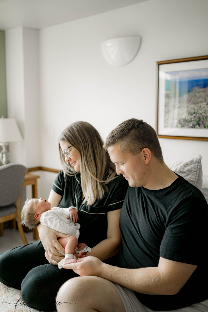 Family cuddling newborn in hospital room – Mom and dad sitting close together, lovingly holding their newborn baby while smiling down at him.