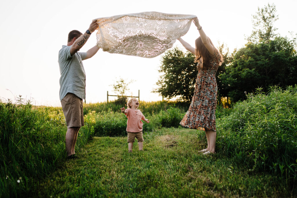 Parents holding lace blanket above toddler as he laughs beneath it during playful family photo.
