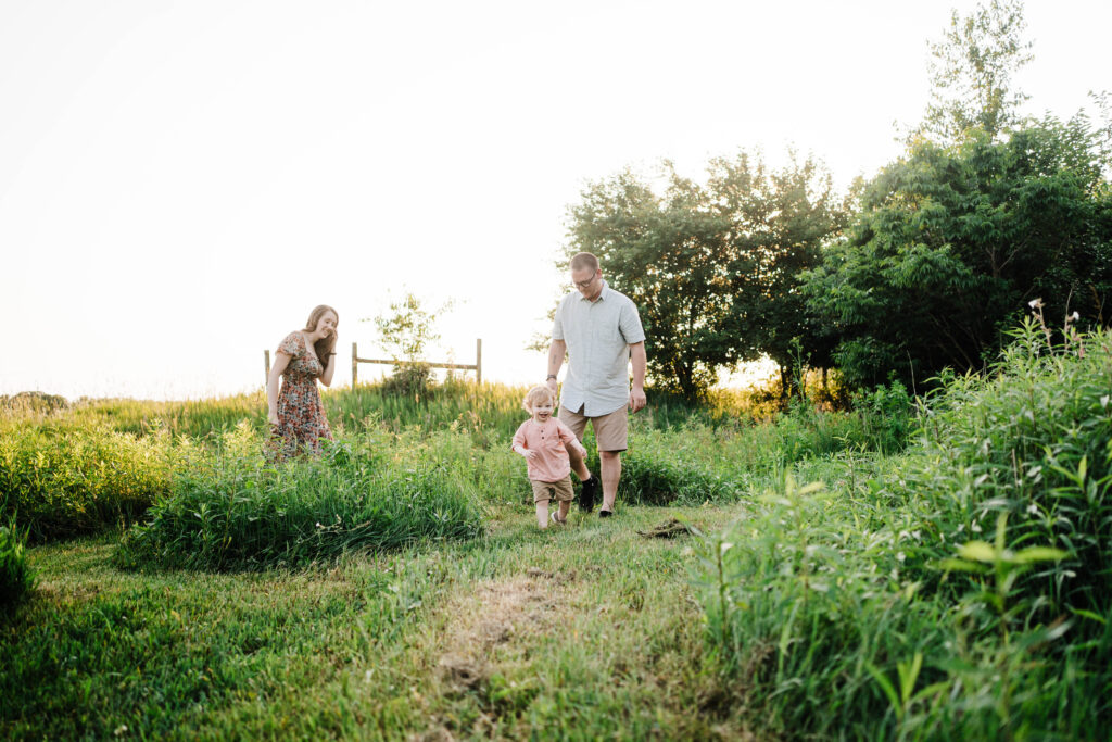 Toddler running toward the camera with parents walking behind in grassy field.