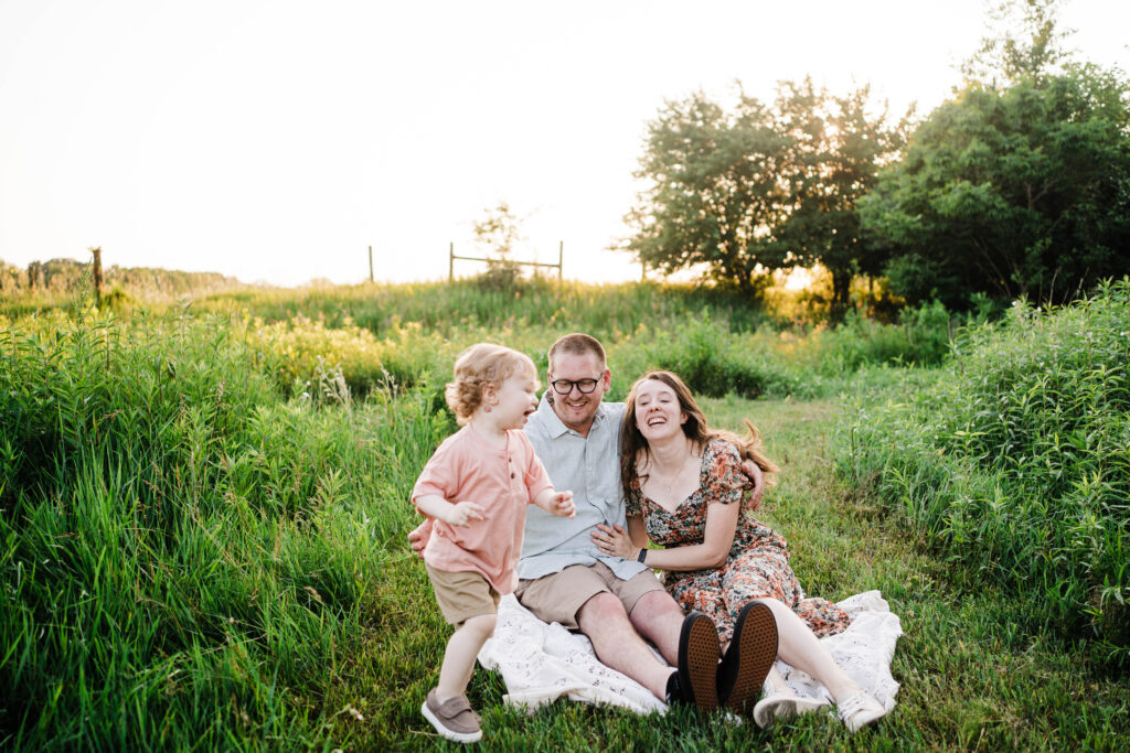 Parents sitting on a blanket in a field laughing together while toddler runs toward them.