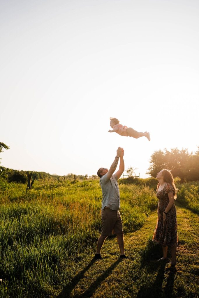 Dad tossing toddler into the air as mom watches and smiles during golden hour family photos.