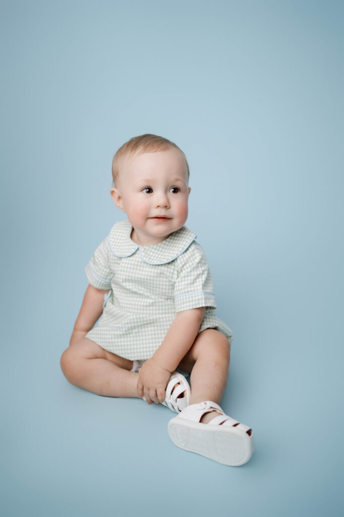 one year old boy in green and white checkered print on a blue backdrop for one year cake portraits.