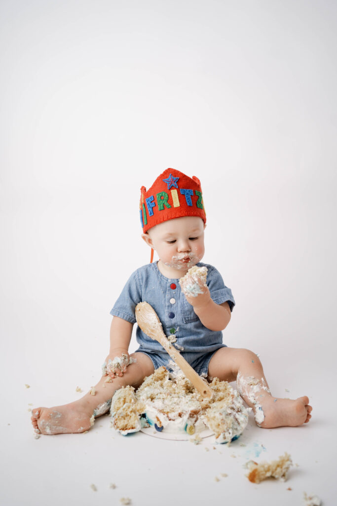 one year old wearing a denim outfit with a red crown while eating cake in a rio, wi portrait studio.