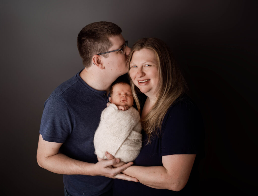 dad kissing mom's temple while they hold their newborn son in a rio, wi studio