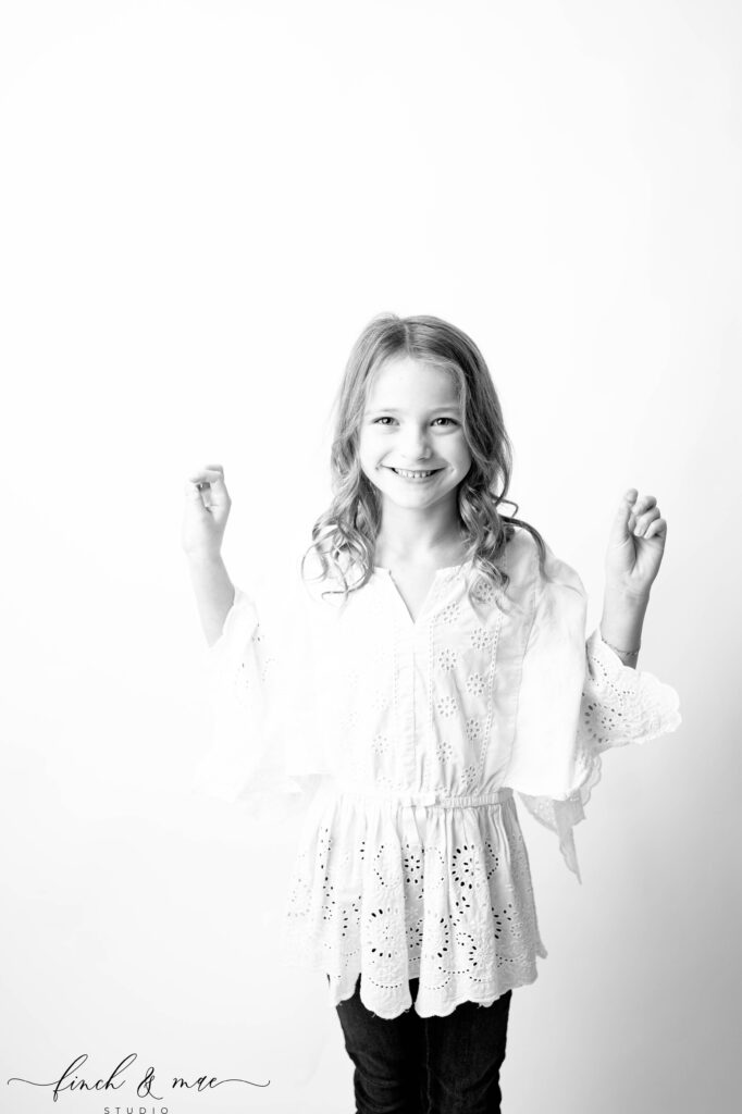 Expressive studio pose of a girl raising her hands playfully, candid children’s portrait session at Finch & Mae Photography.