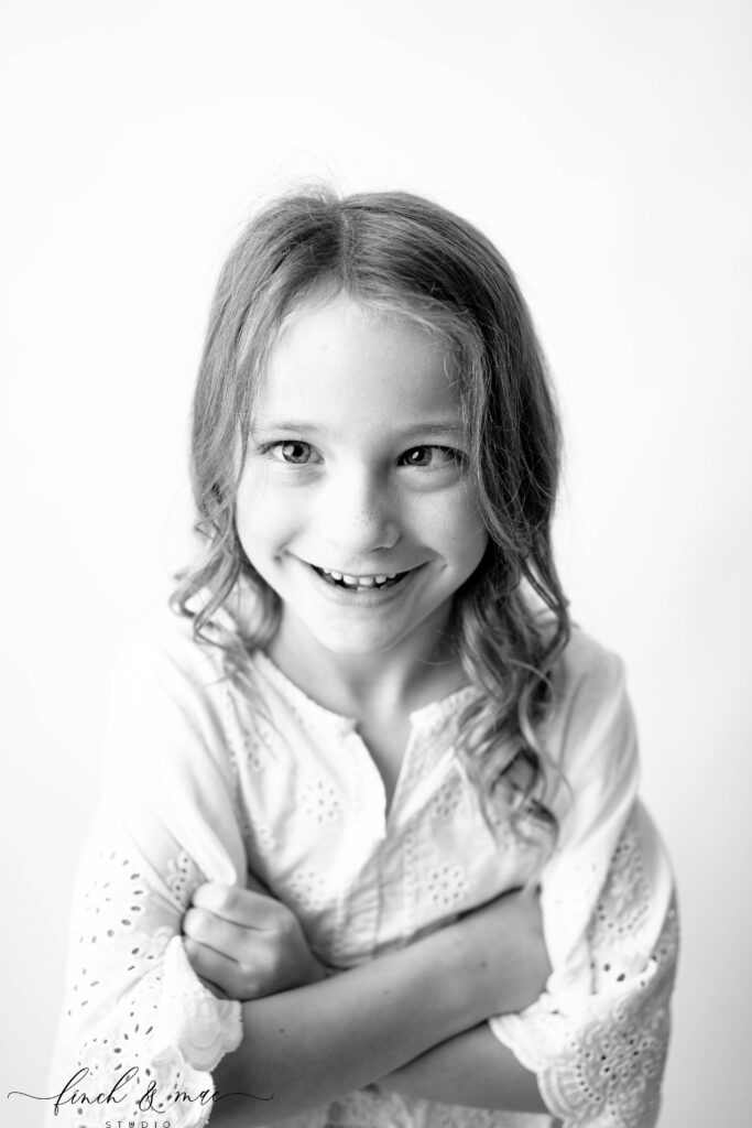 Black and white studio portrait of a young girl smiling with crossed arms, captured naturally by Wisconsin child photographer Finch & Mae Studio.