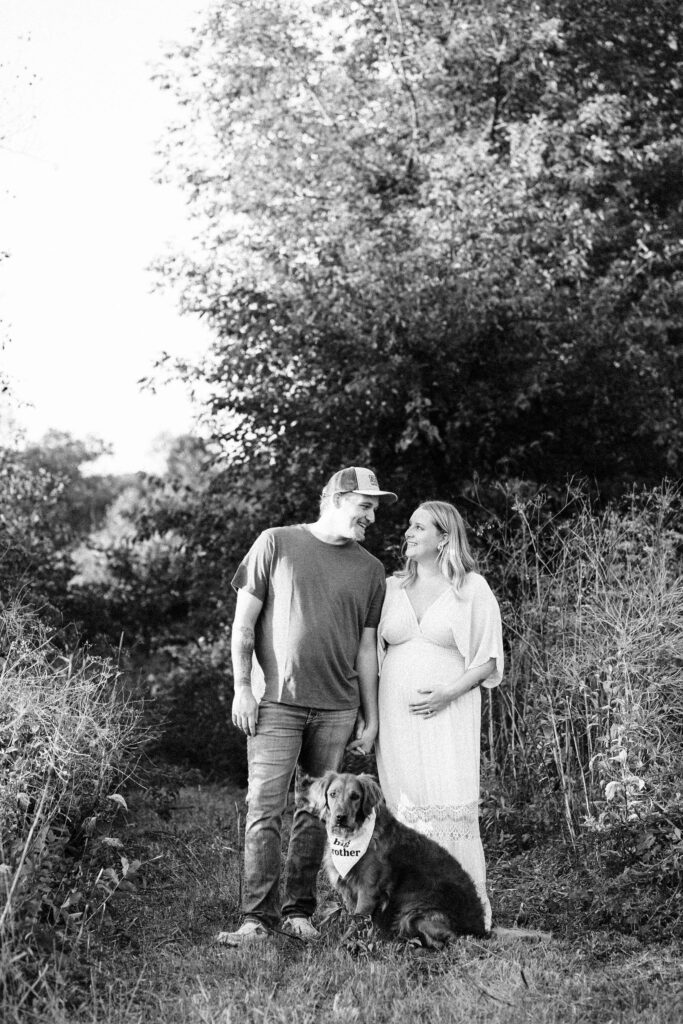 Black and white photo of a couple, staring into each other’s eyes while Mama Carissa is expecting belly and the golden retriever wears a big brother bandanna