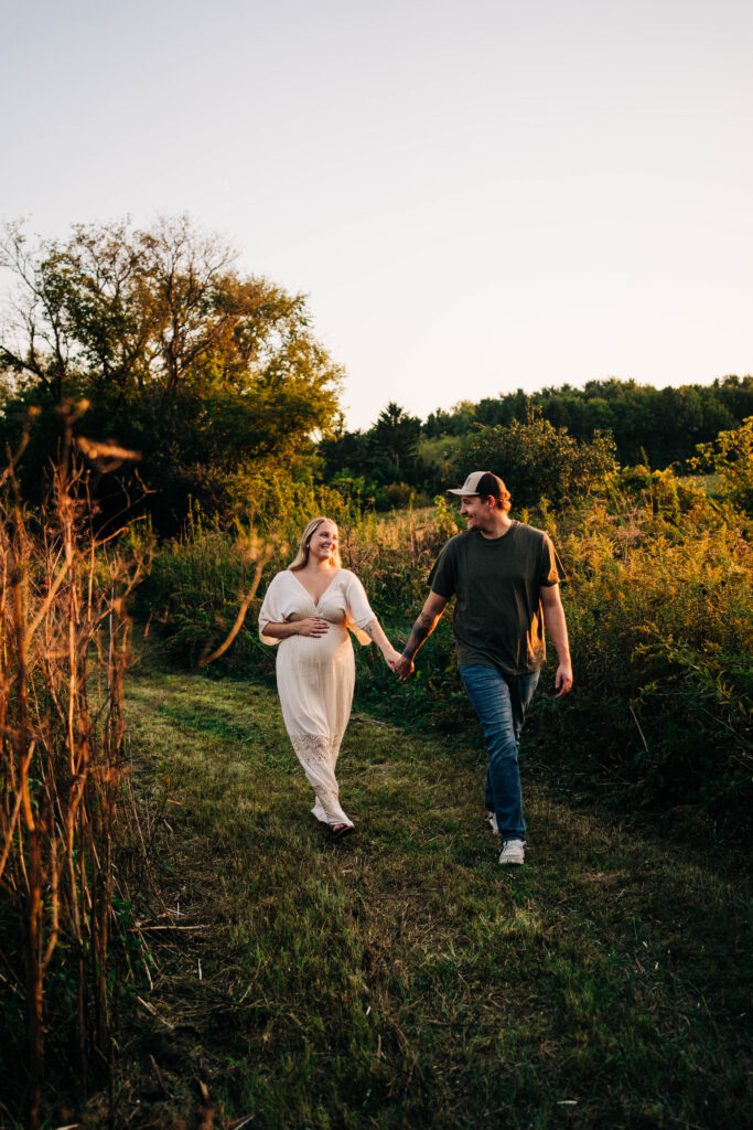 An expecting couple is holding hands and walking towards the camera in Sun, Prairie, Wisconsin