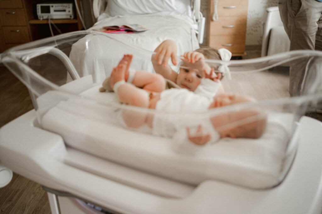 Big sister reaching into hospital bassinet to touch baby brother at aurora medical center in wi