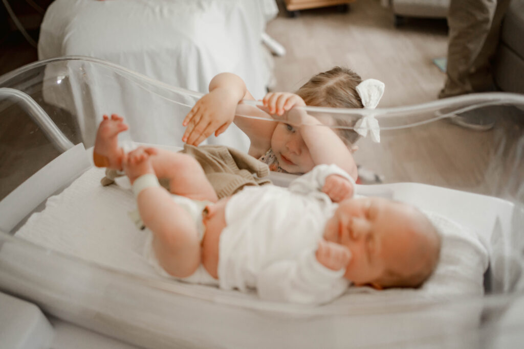Big sister reaching into hospital bassinet to touch baby brother during Madison fresh 48 hospital session