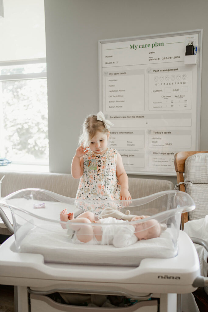 Big sister looking into the hospital bassinet at her baby brother for a fresh 48 session at auroral medical center in wisconsin