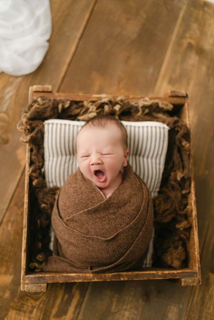 yawning newborn boy wrapped in brown wrap in a wooden box - newborn portrait studio near sun prairie, wi