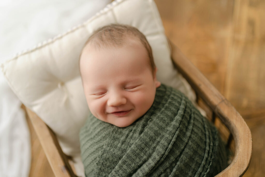 newborn boy in a green wrap is smiling in newborn portrait studio