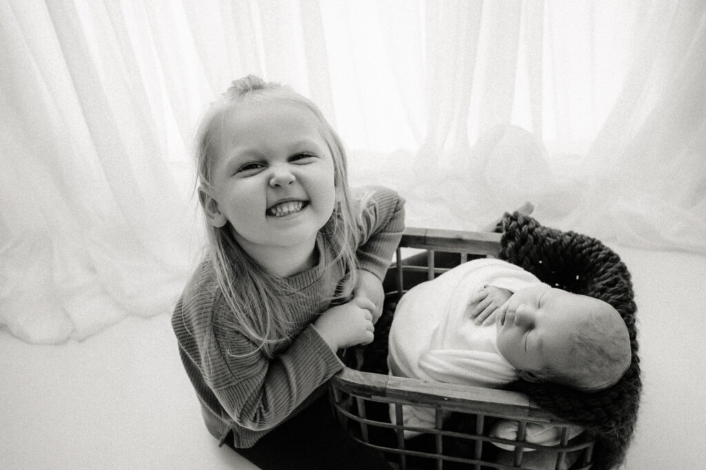 Big sister smiling with newborn brother in a wooden basket - Newborn photography session near Madison, WI